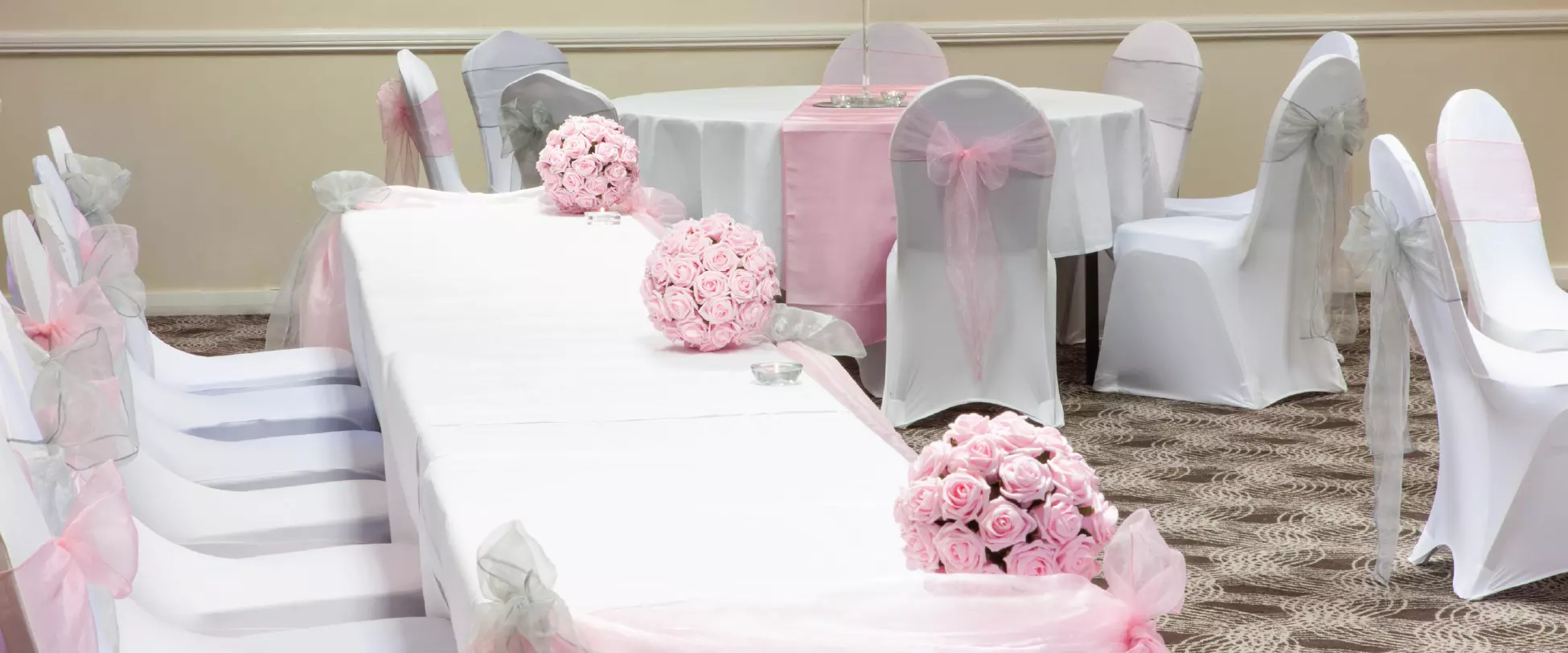 Tables set up for a wedding breakfast with white table covers, chair covers, and pink flowers and ribbons..